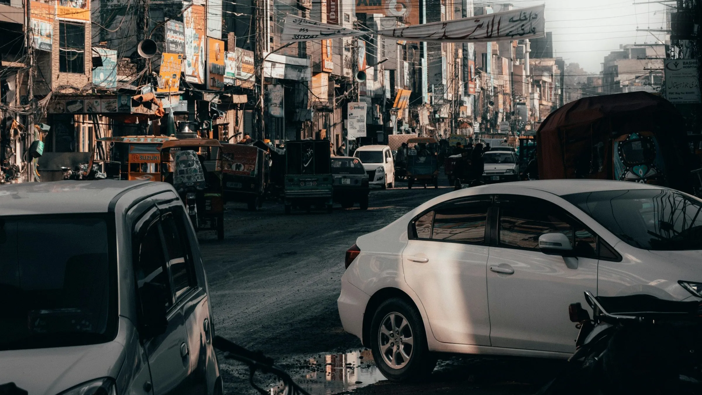 A busy street in Lahore with parked cars, rickshaws, shops and signboards